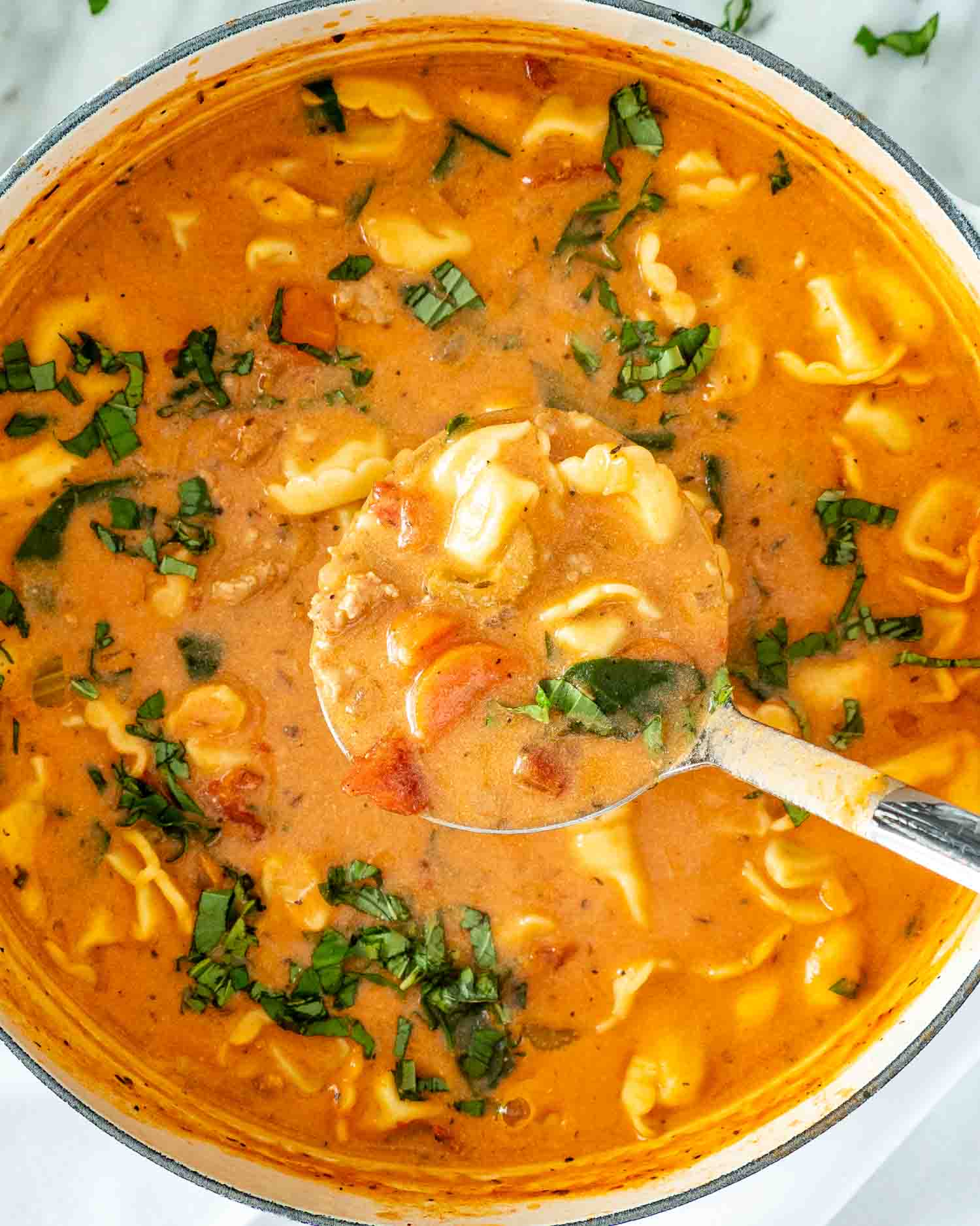 Overhead close-up of a silver ladle filled with creamy sausage tortellini soup, with chopped basil scattered across the pot.
