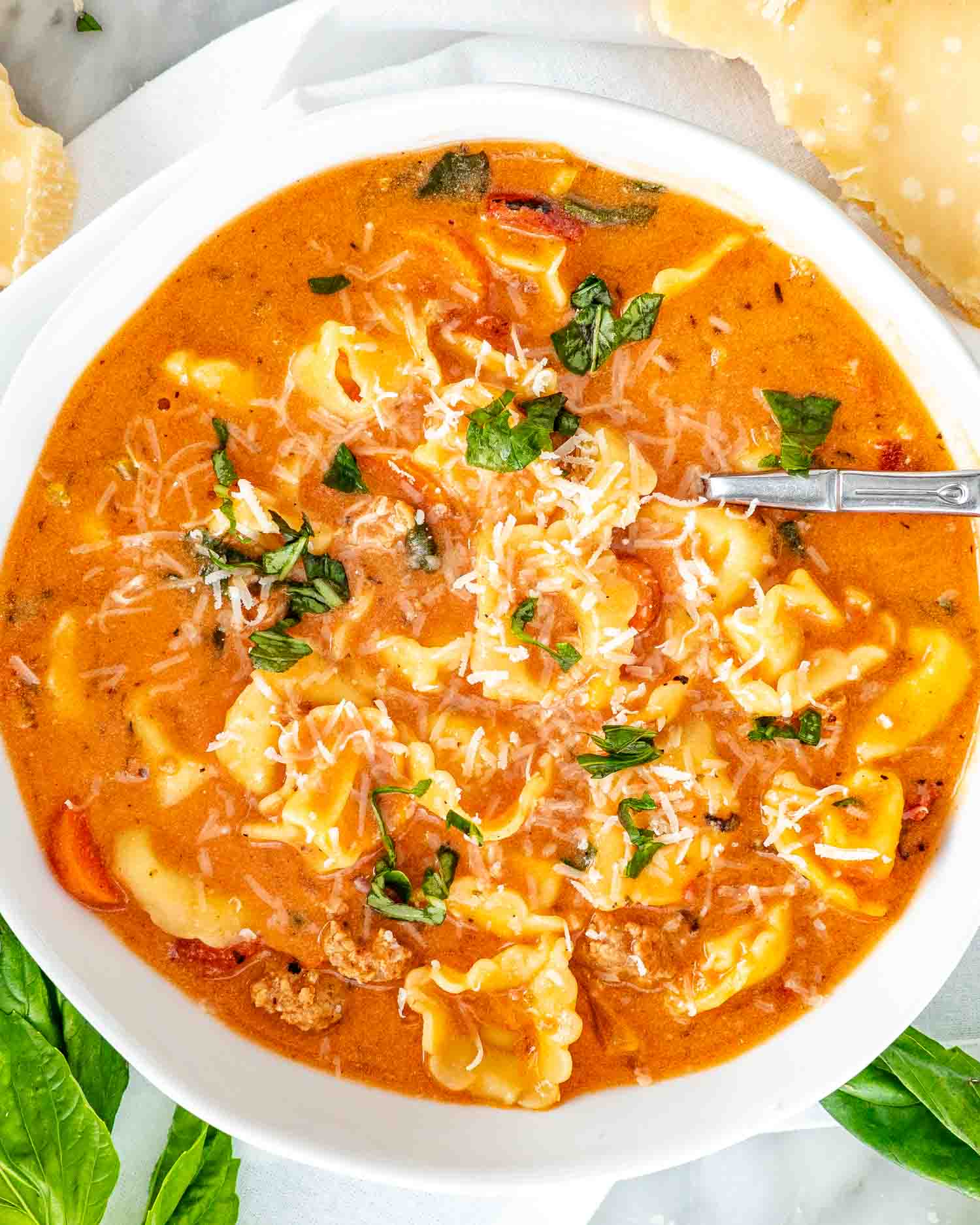 Overhead view of creamy sausage tortellini soup topped with grated Parmesan and fresh basil, served in a white bowl.