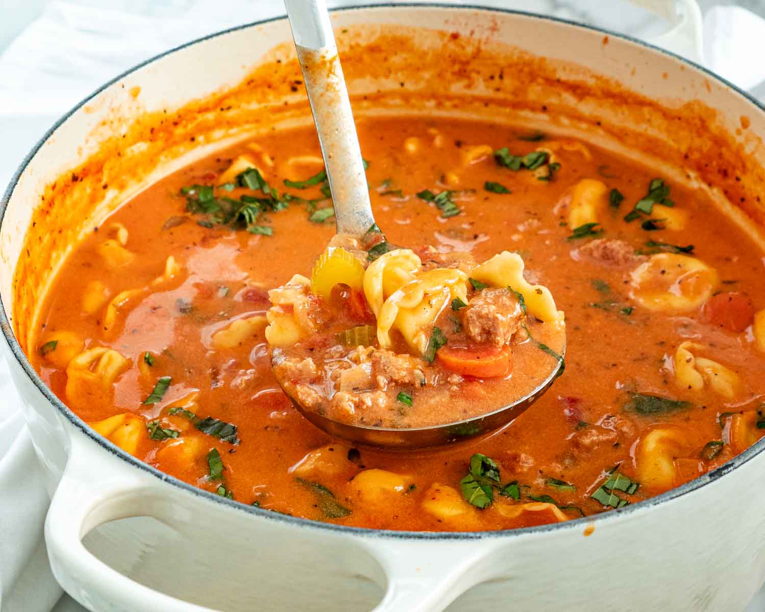 Close view of a ladle holding tortellini, sausage, carrots and celery above creamy tomato soup in a white pot.