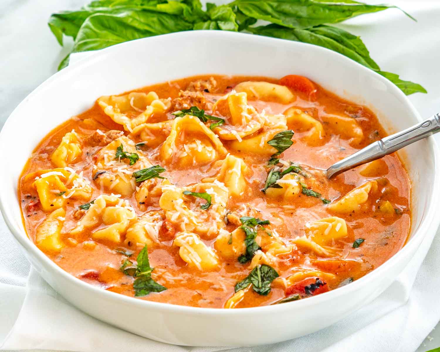 Creamy sausage tortellini soup in a shallow white bowl, sprinkled with grated Parmesan and chopped basil, with a silver spoon.