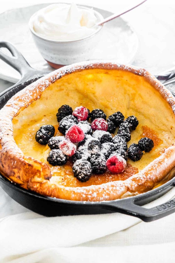 dutch baby pancake in a skillet topped with fresh berries and dusted with powdered sugar. A small bowl of whipped cream is in the background