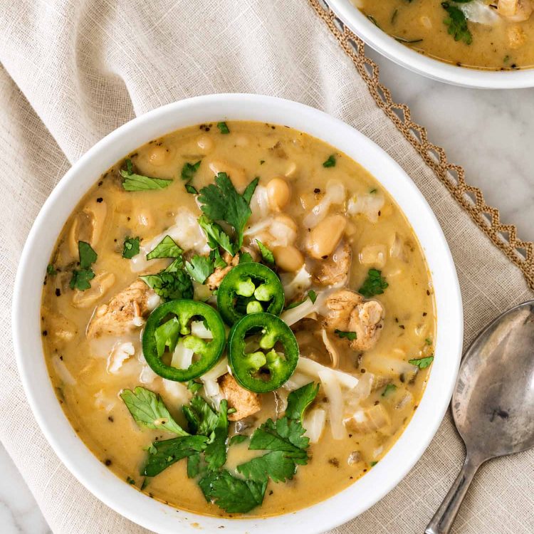 Overhead view of white chicken chili topped with jalapeño slices, shredded cheese, and cilantro, served on beige linen.