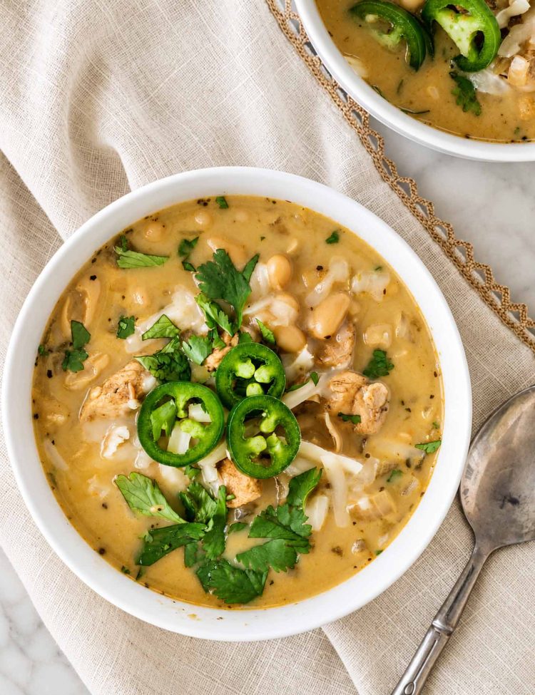 Overhead view of white chicken chili topped with jalapeรฑo slices, shredded cheese, and cilantro, served on beige linen.