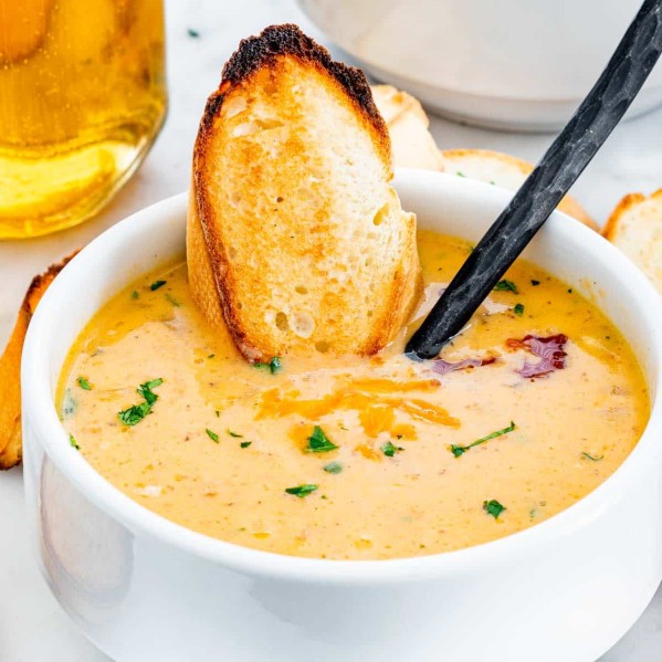 a bowl of bacon and beer cheese soup with a piece of toasted baguette and a spoon in it