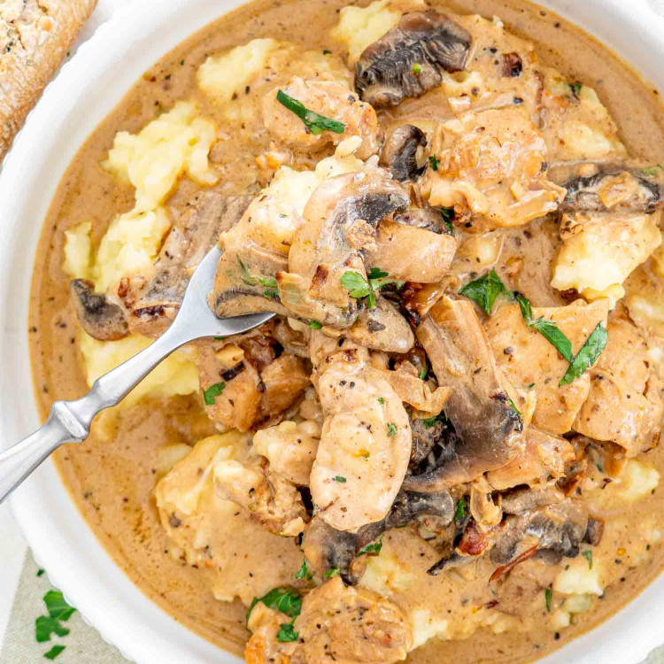 A bowl of chicken stroganoff with mushrooms atop smooth mashed potatoes, sprinkled with herbs.