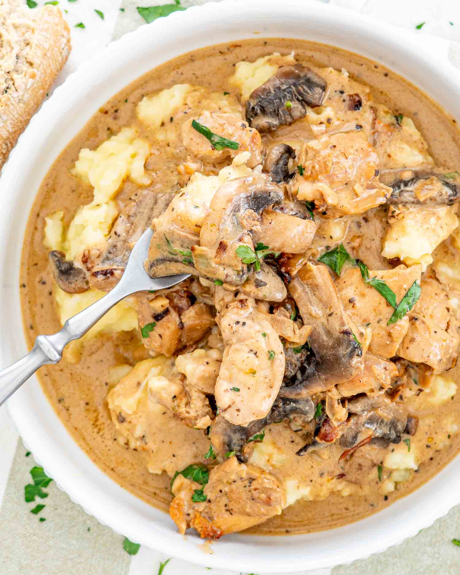 A bowl of chicken stroganoff with mushrooms atop smooth mashed potatoes, sprinkled with herbs.