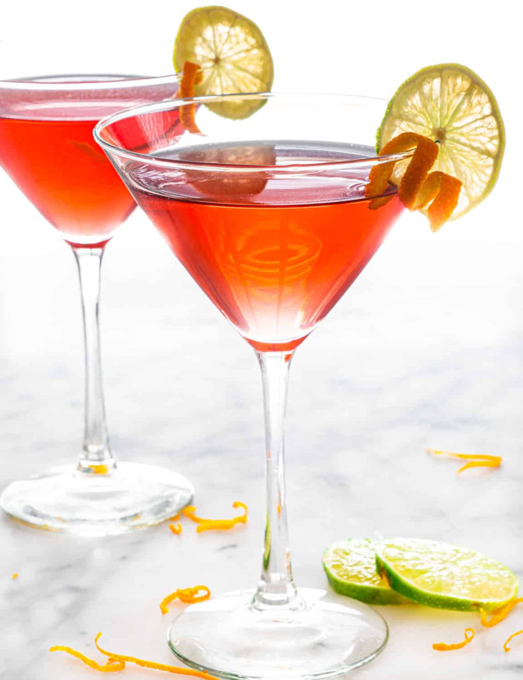 two cosmopolitan cocktails in martini glasses garnished with orange twists and lime slices