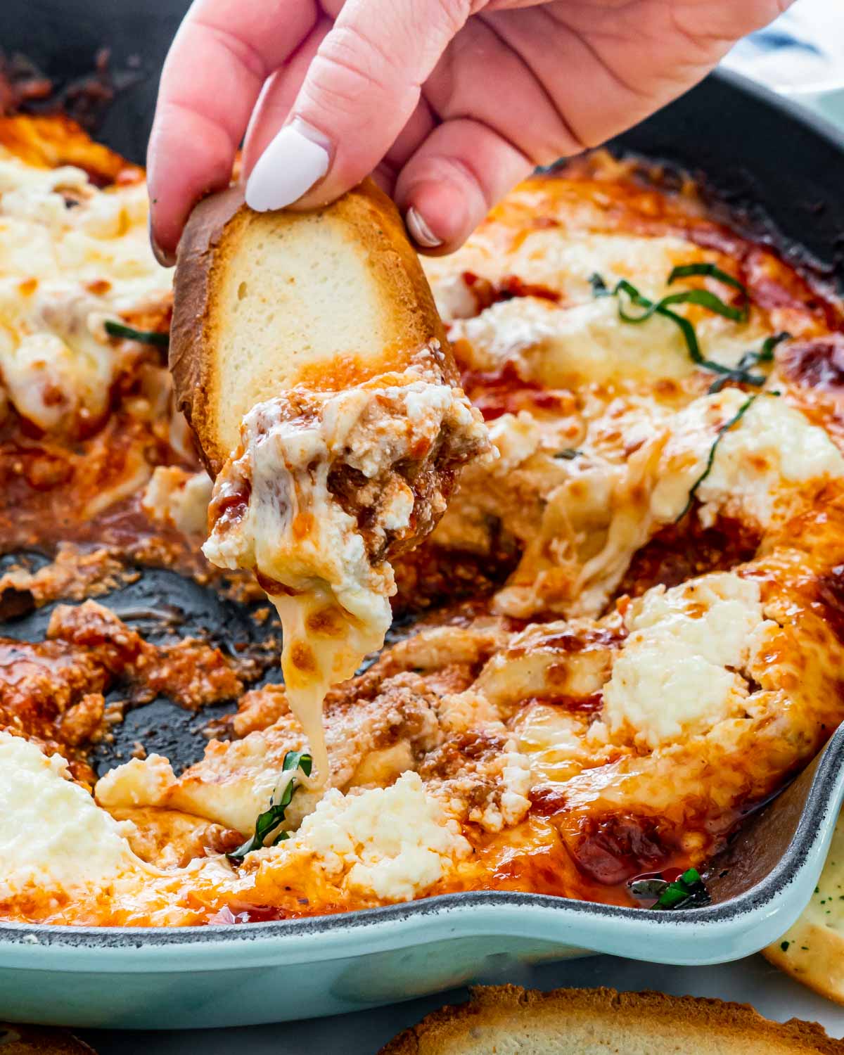 a hand dipping a piece of toast in a skillet with lasagna dip