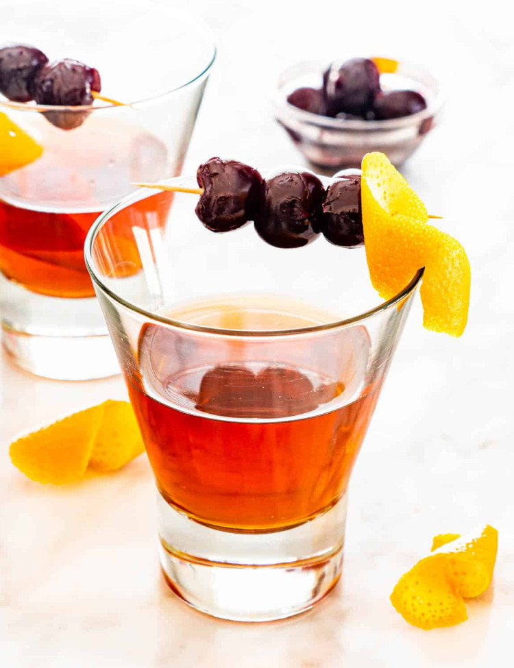a manhattan drink in a glass garnished with an orange twist and bourbon cherries