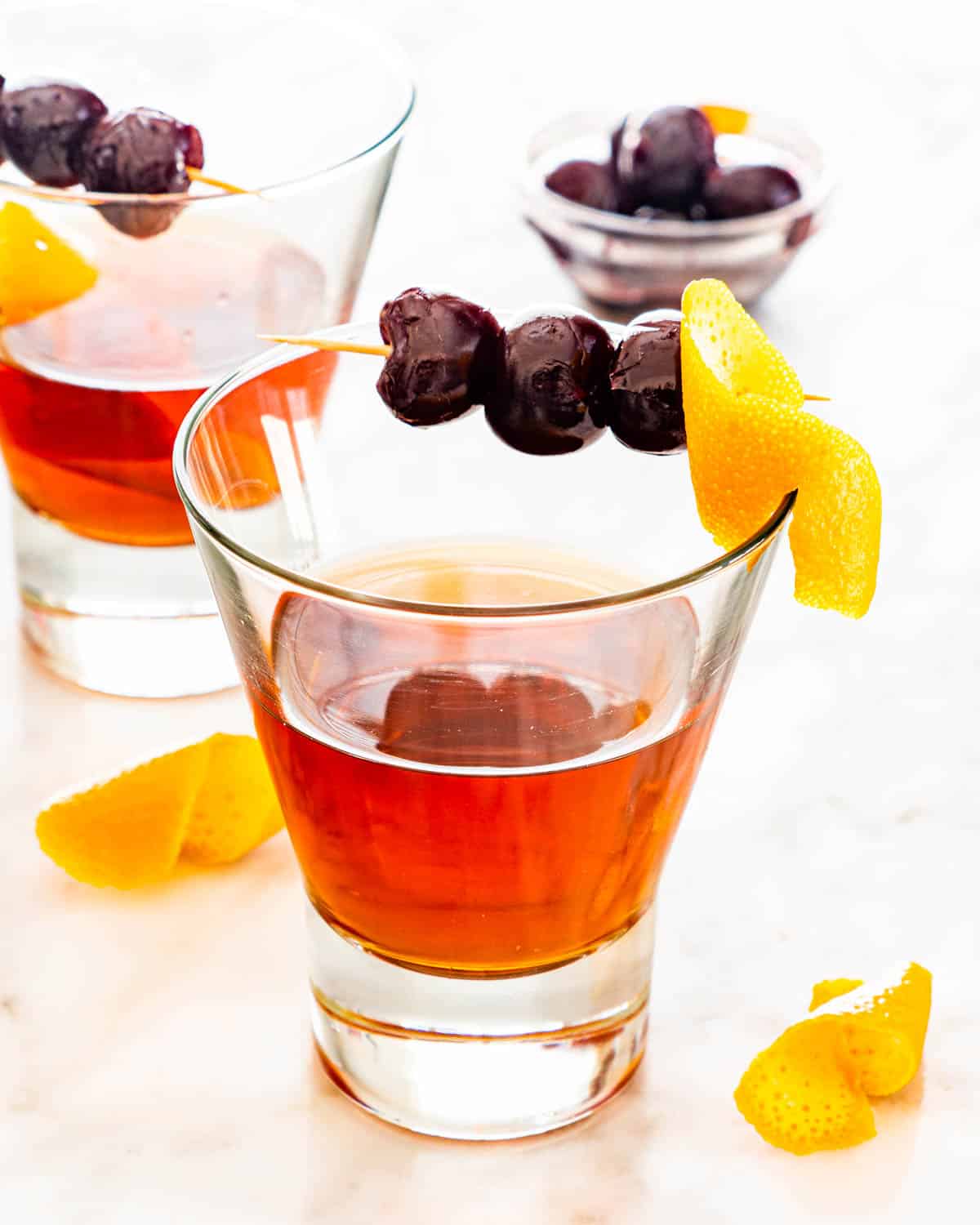 two glasses with manhattan garnished with cherries and ornage peel