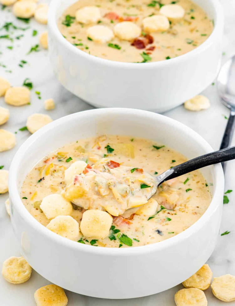 side view shot of a spoon taking a bite of new england clam chowder from a bowl surrounded with oyster crackers