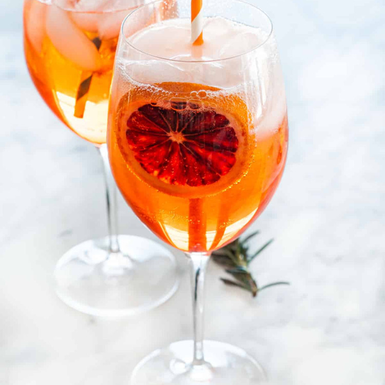 two wine glasses full of aperol spritz garnished with a slice of blood orange and straw