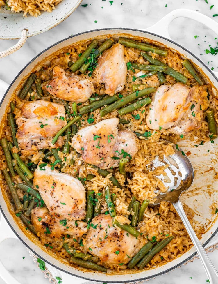 overhead shot of coconut chicken with rice and green beans in a large beige braiser with some rice already taken out and a serving spoon inside