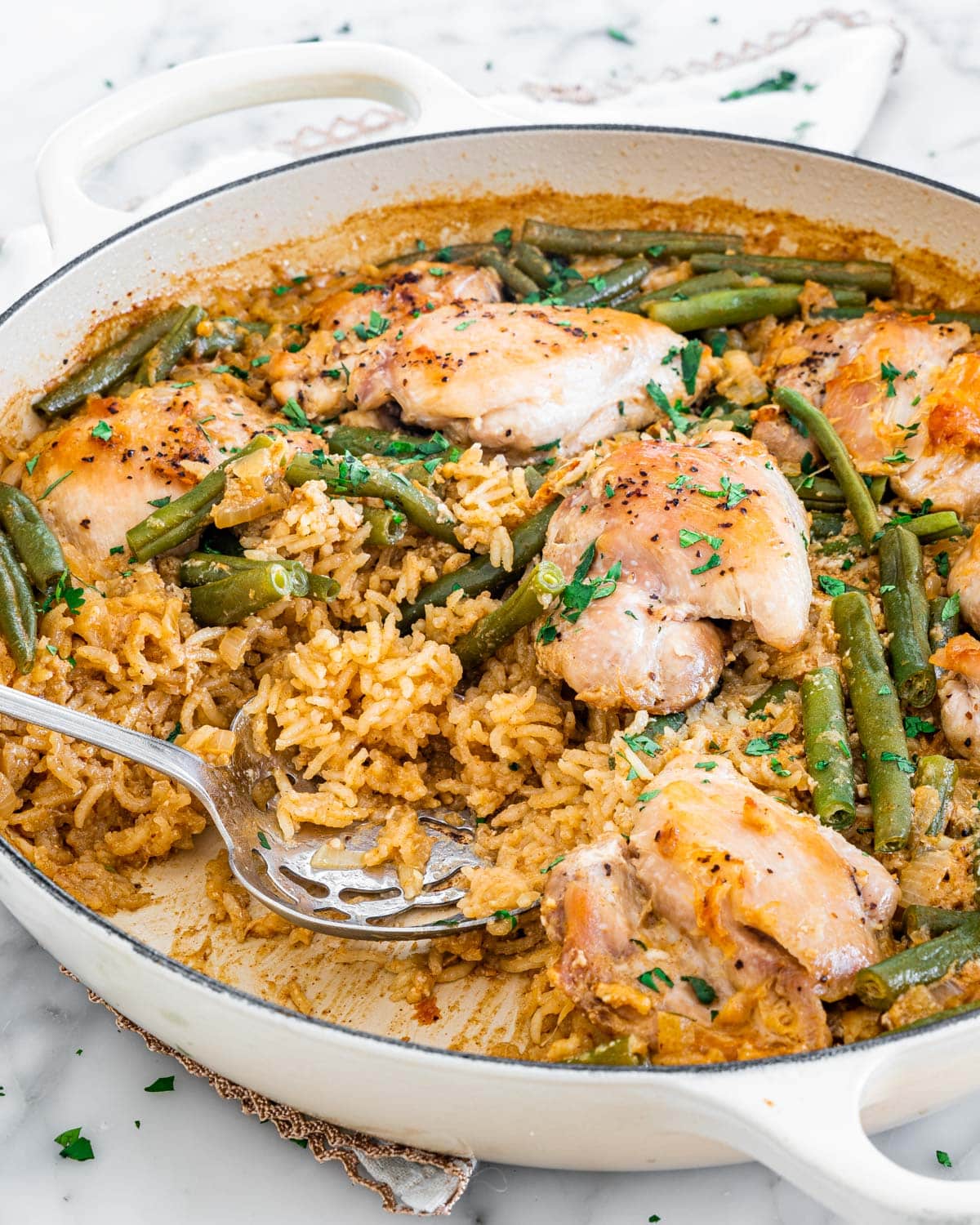 side shot of coconut chicken with rice and green beans in a beige braiser garnished with cilantro and with a serving spoon inside