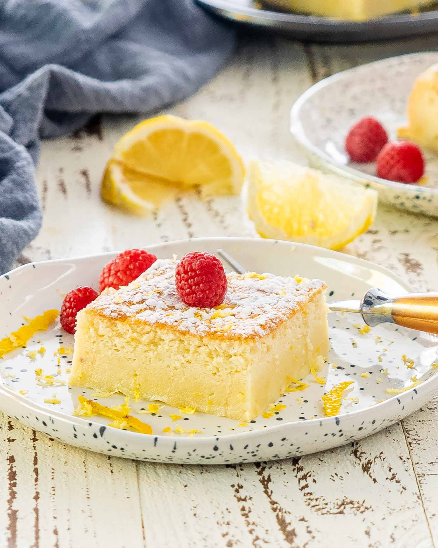 a slice of lemon magic cake on a white plate with a raspberry on top.