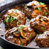 side view shot of salisbury steaks in a skillet, topped with mushrooms, garnished with chopped parsley