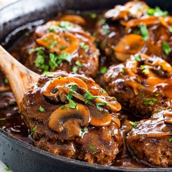 a wooden spoon lifting a salisbury steak out of the skillet