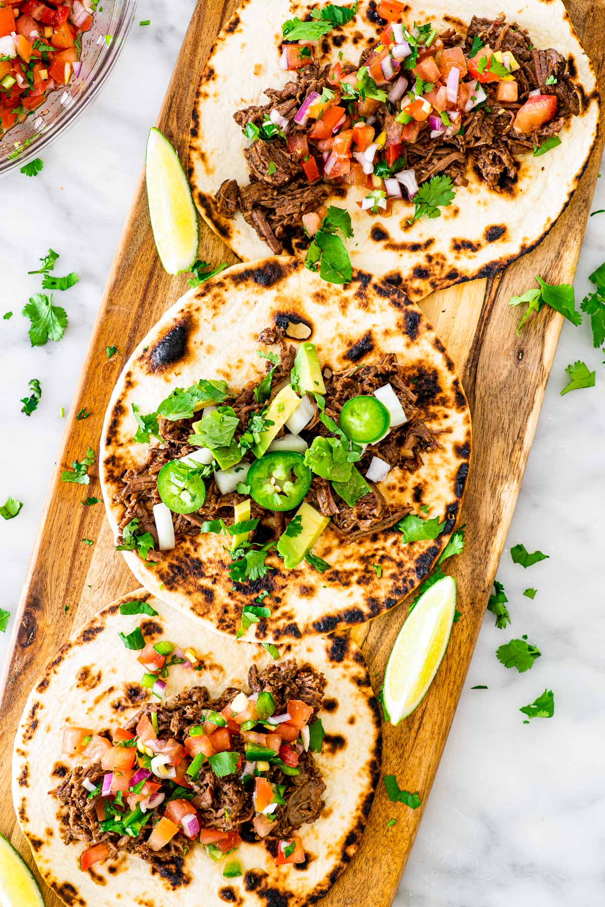 overhead shot of 3 tacos on a long cutting board with barbacoa beef, some topped with pico de gallo and some with onion, avocado and jalapeno peppers