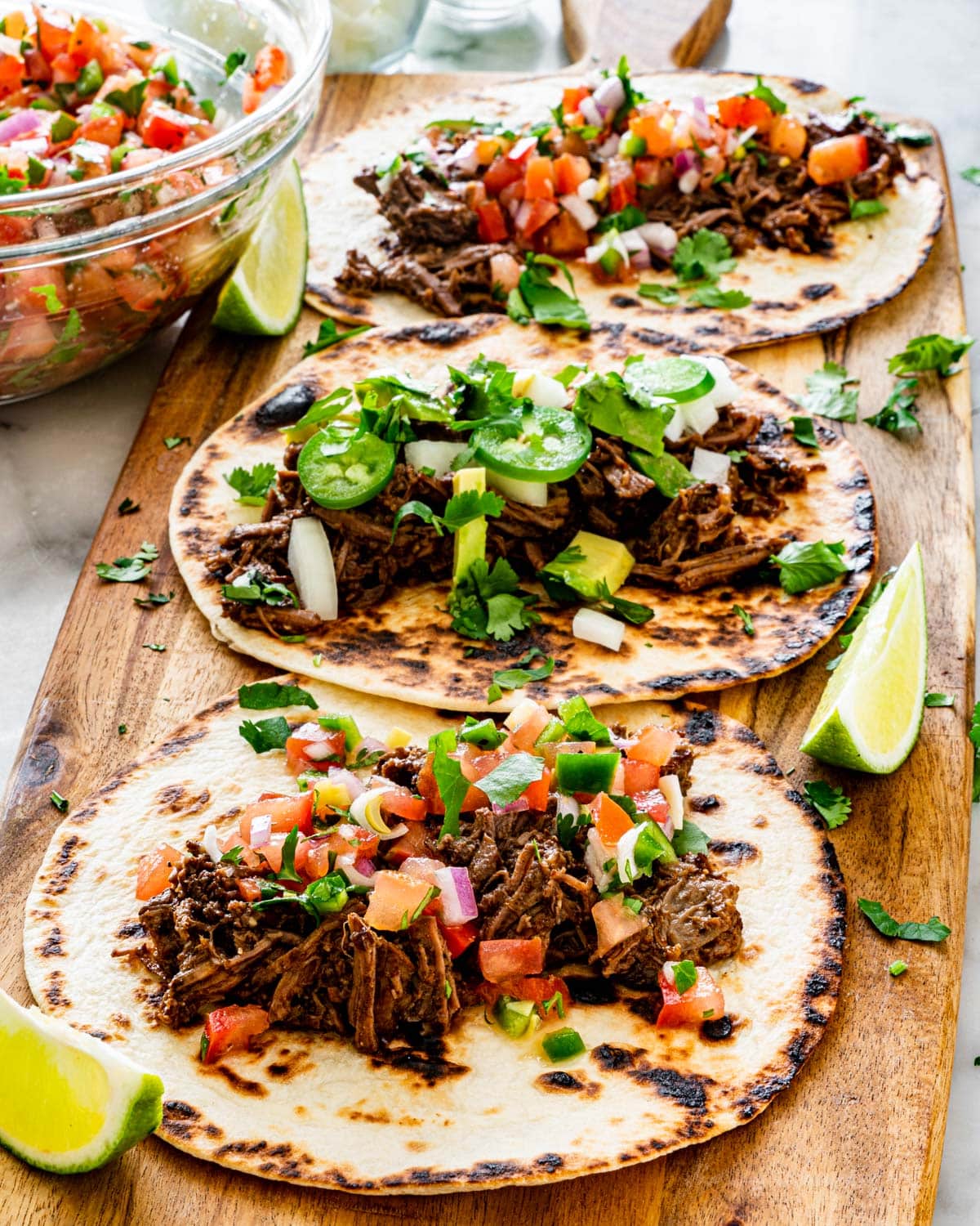 side shot shot of 3 tacos on a long cutting board with barbacoa beef, some topped with pico de gallo and some with onion, avocado and jalapeno peppers