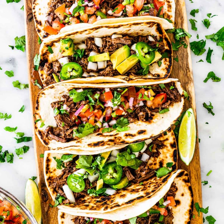 overhead shot of a bunch of tacos on a long cutting board with barbacoa beef, some topped with pico de gallo and some with onion, avocado and jalapeno peppers