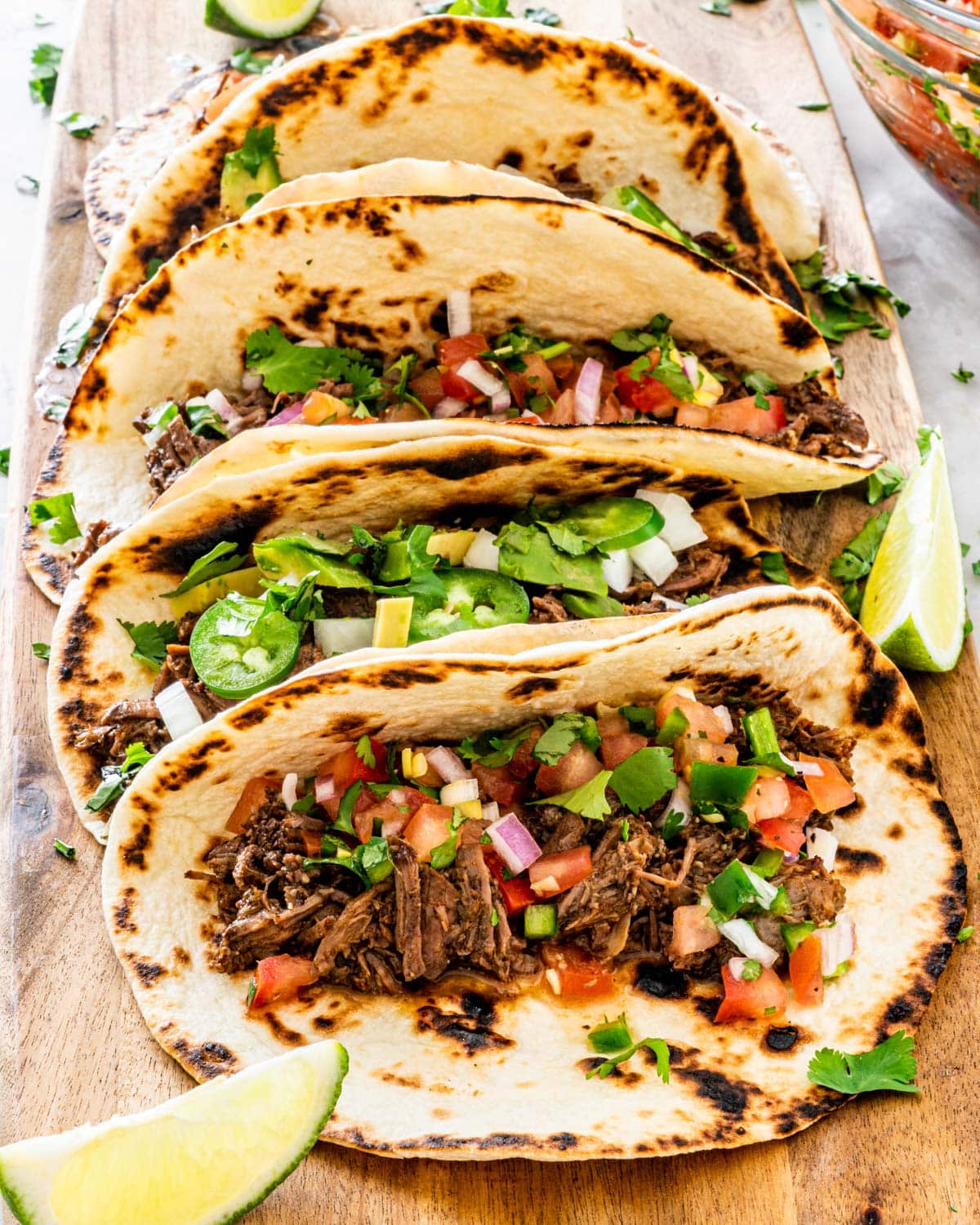 side shot of a bunch of tacos on a long cutting board with barbacoa beef, some topped with pico de gallo and some with onion, avocado and jalapeno peppers