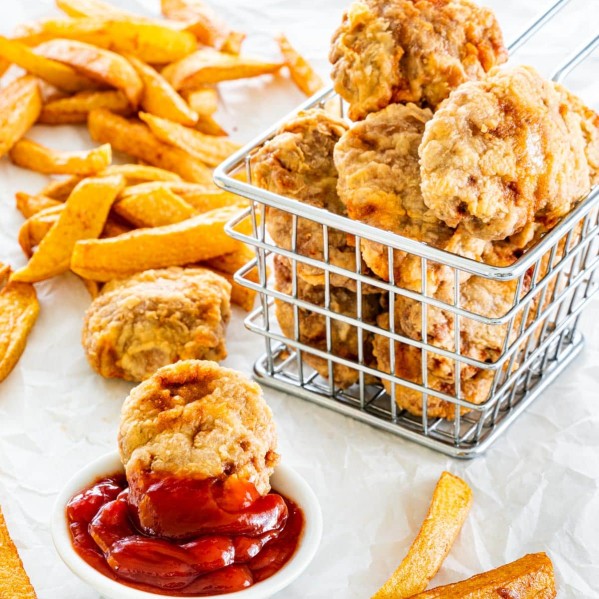 side picture of a basket full of chicken nuggets with a nugget in a small bowl with ketchup and fries in the background