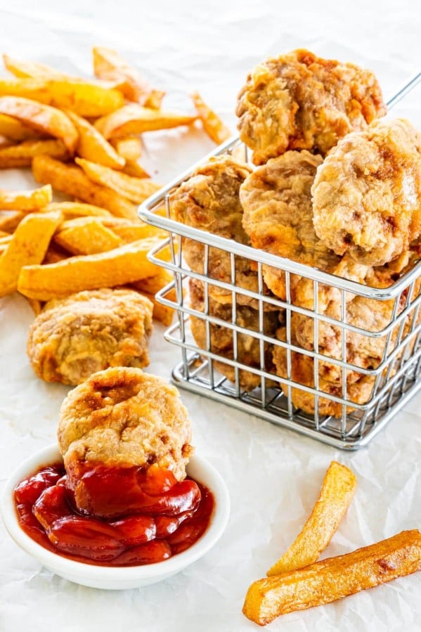 side picture of a basket full of chicken nuggets with a nugget in a small bowl with ketchup and fries in the background