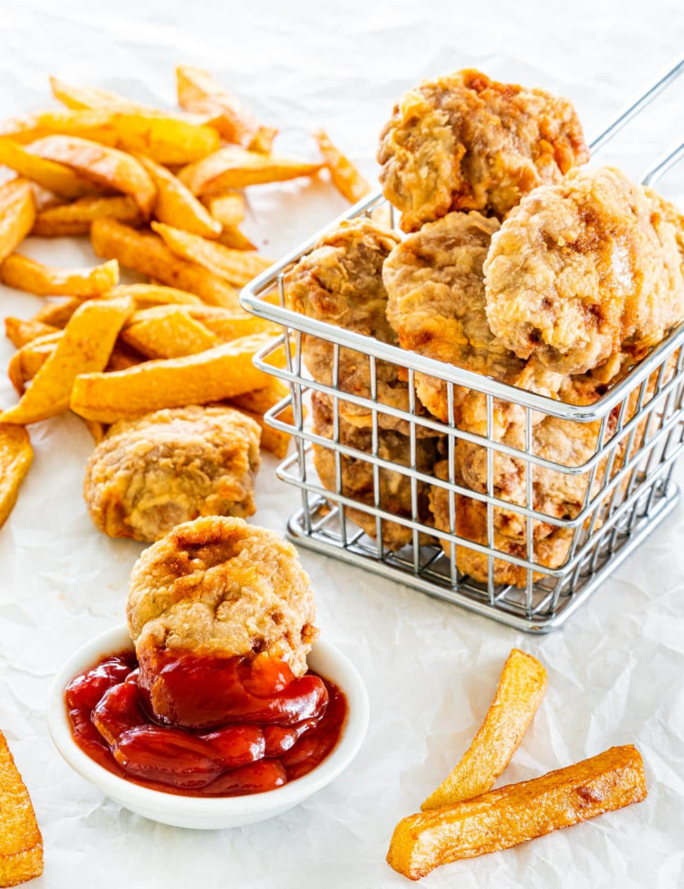 side picture of a basket full of chicken nuggets with a nugget in a small bowl with ketchup and fries in the background