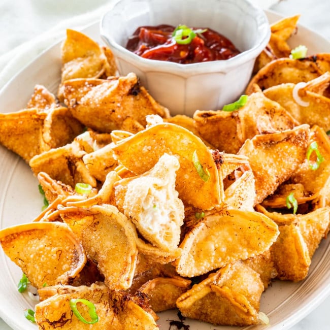 side view shot of crab rangoon on a serving platter with sweet and sour sauce in a little white bowl