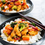 side shot of two black plates with hunan chicken over a bed of rice with chop sticks on top of the plates