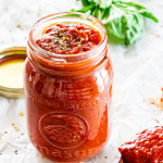 side view shot of freshly homemade pizza sauce in a jar with some basil in the background and a mini wooden spoon with some sauce next to the jar