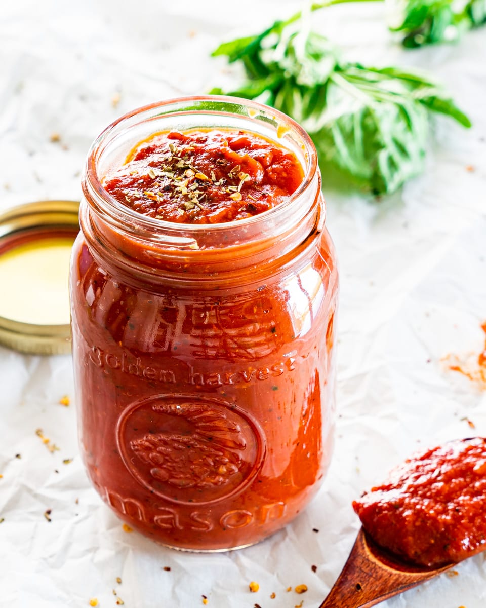side view shot of freshly homemade pizza sauce in a jar with some basil in the background and a mini wooden spoon with some sauce next to the jar