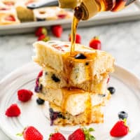 a hand pouring maple syrup over a stack of pancakes surrounded by berries