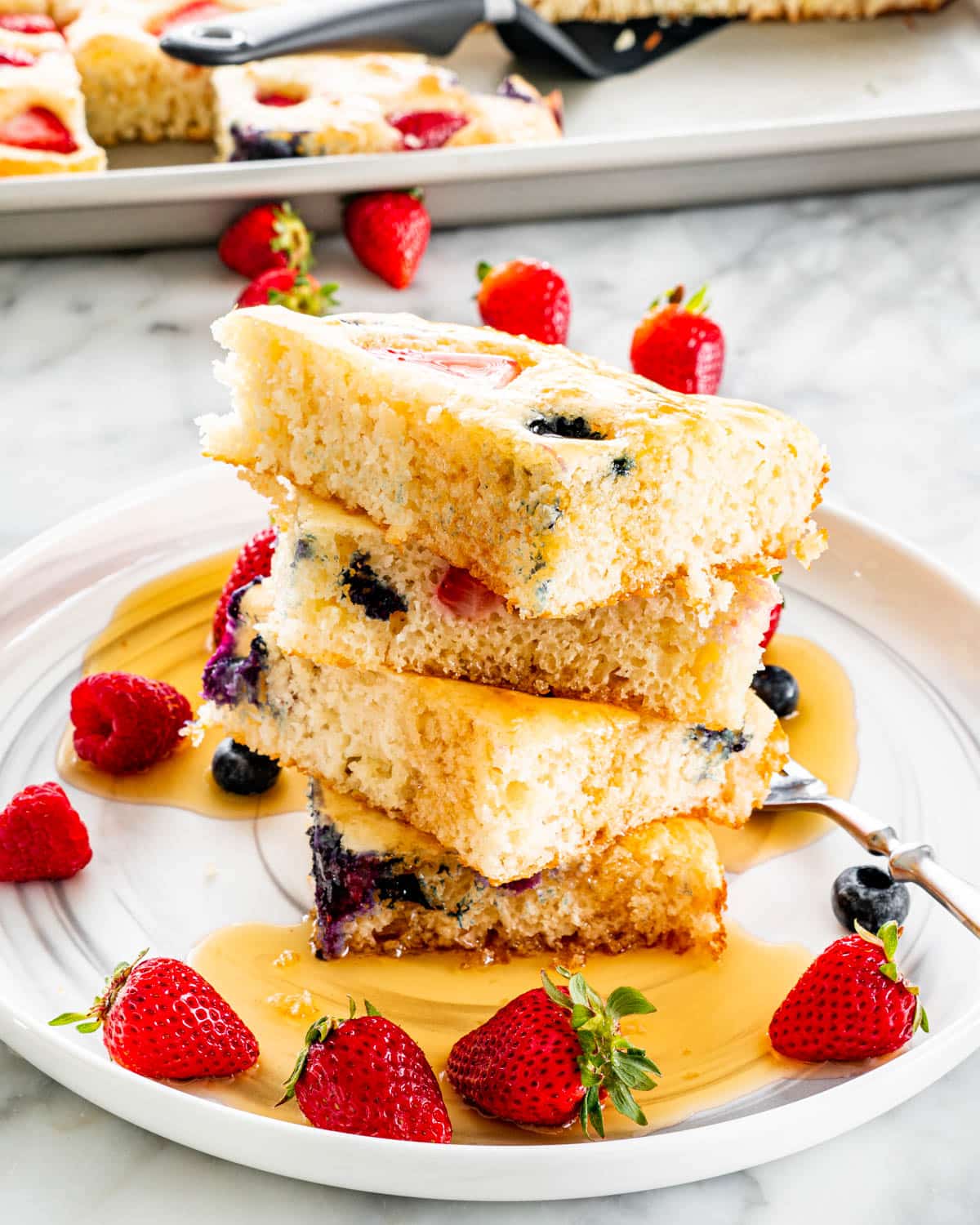 a stack of sliced up sheet pan pancakes on a white plate drizzled with maple syrup and lots of berries