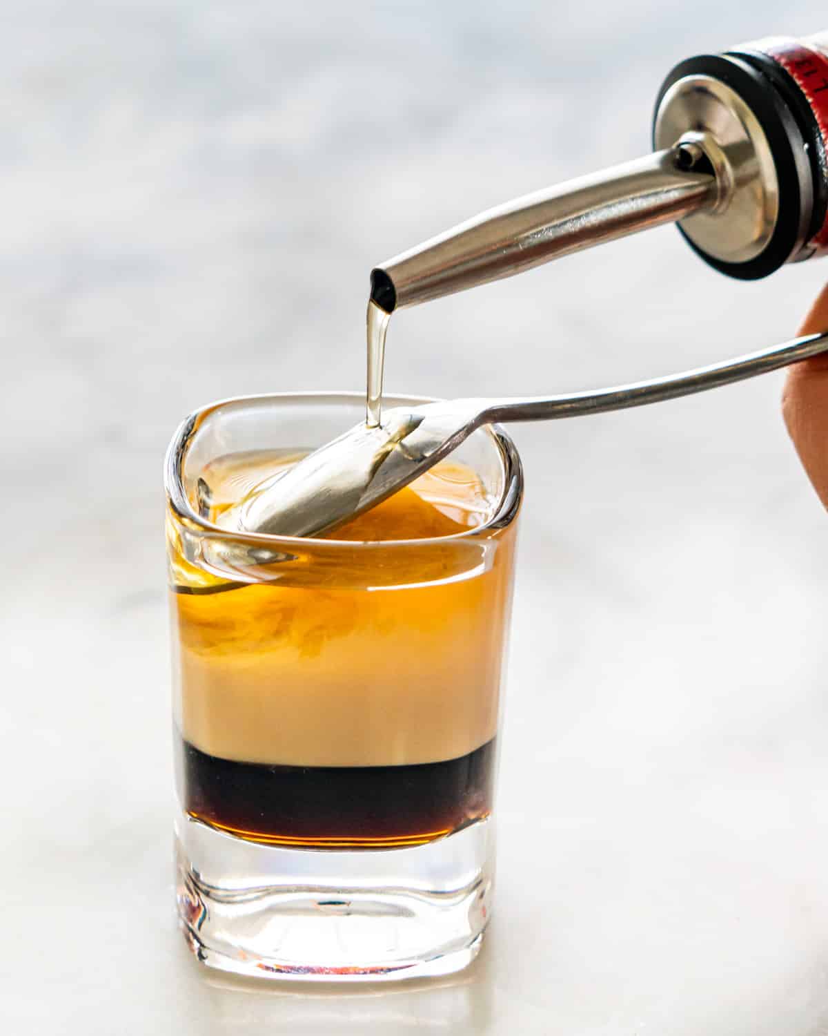 side view shot of pouring grand marnier over a teaspoon in a shot glass with 2 layers one of coffee liqueur and one of irish cream
