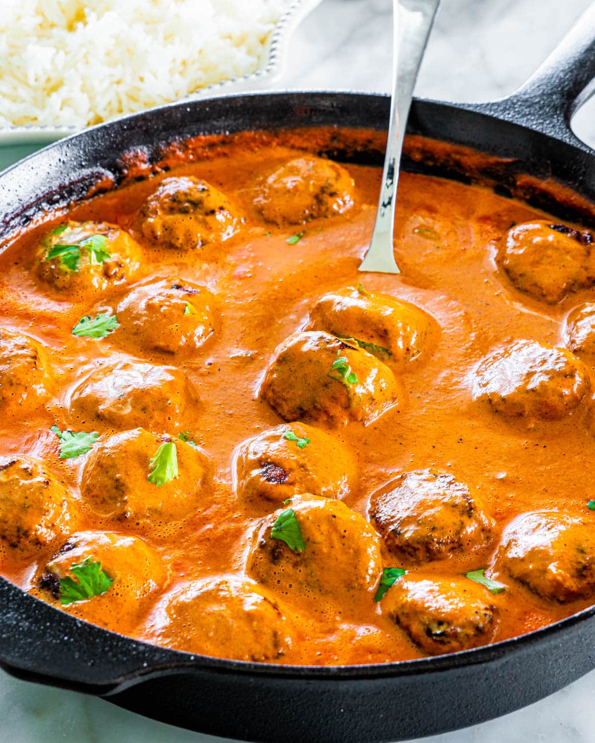 side shot of butter chicken meatballs in a black skillet with a serving spoon inside