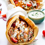 a chicken gyro wrapped in paper and topped with lots of tzatziki sauce with another gyro in the background