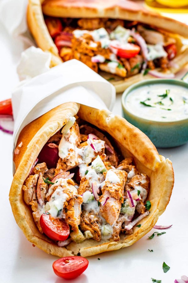 a chicken gyro wrapped in paper and topped with lots of tzatziki sauce with another gyro in the background