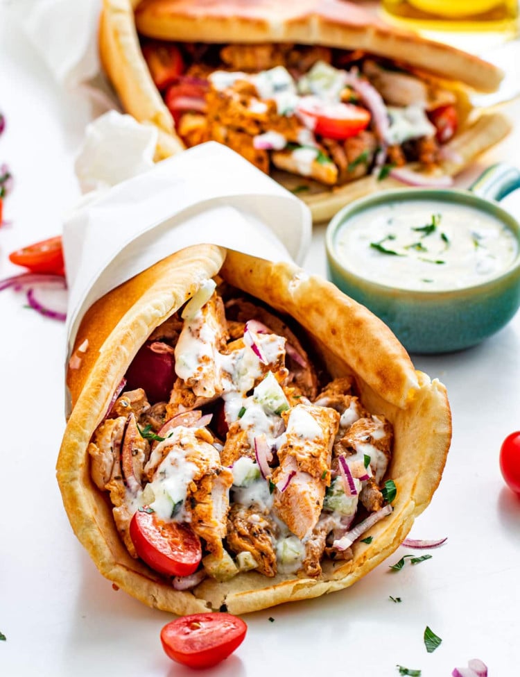 a chicken gyro wrapped in paper and topped with lots of tzatziki sauce with another gyro in the background
