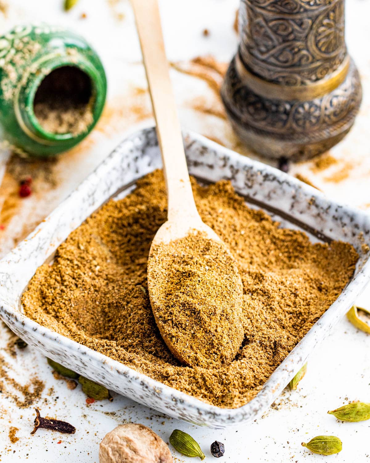 a square bowl filled with garam masala and a mini wooden spoon inside