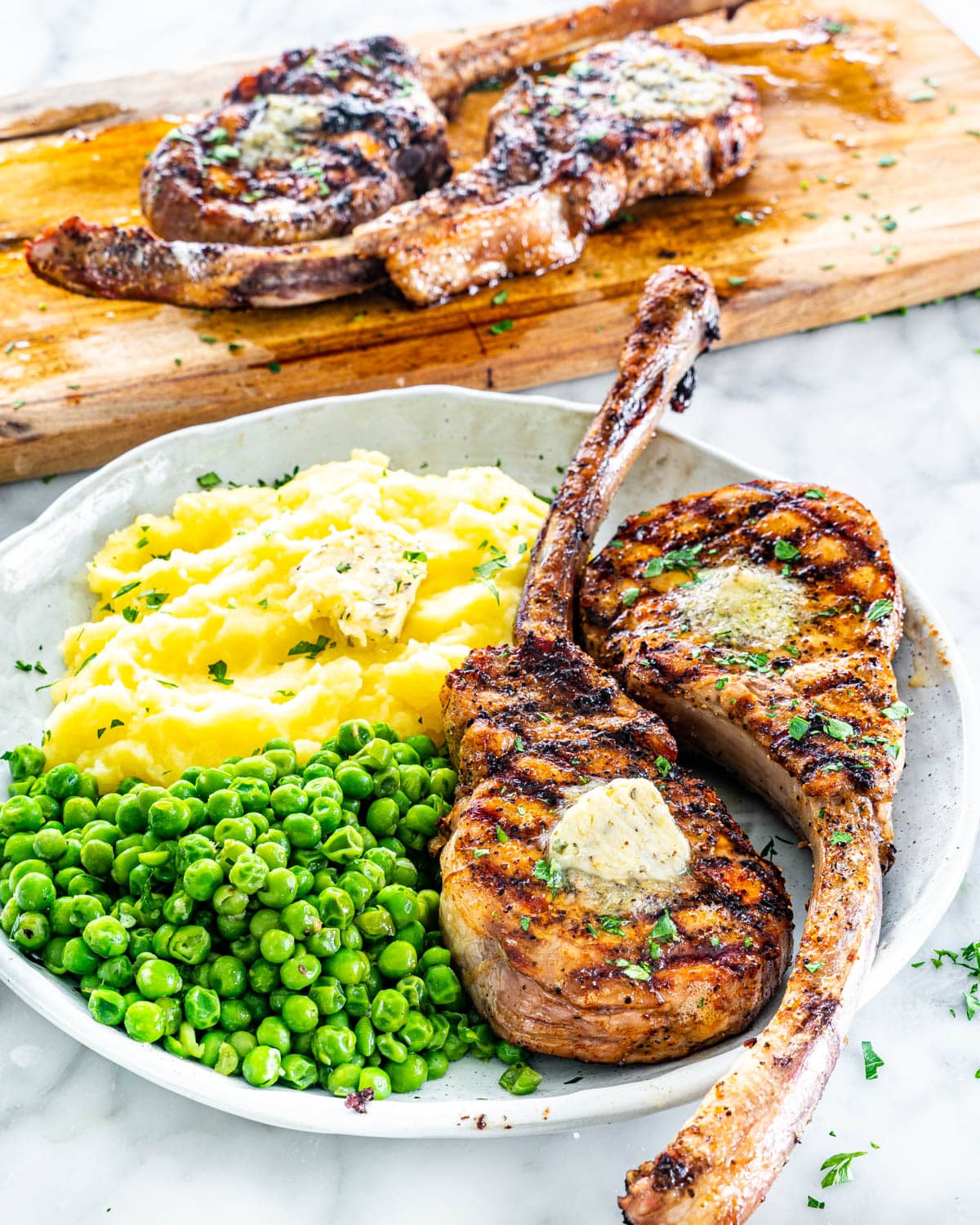 side view shot of two tomahawk grilled pork chops on a plate with mashed potatoes and peas