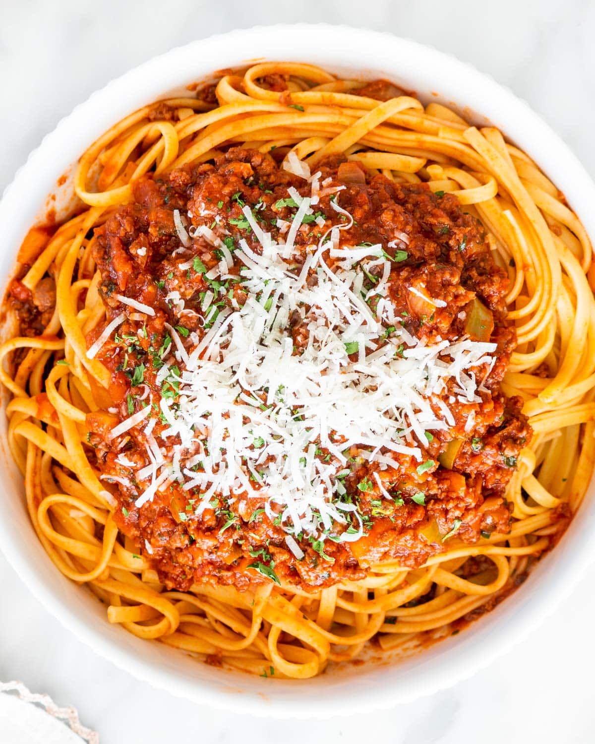 overhead shot of linguini in a white bowl with lots of meat sauce and garnished with parmesan cheese and parsley