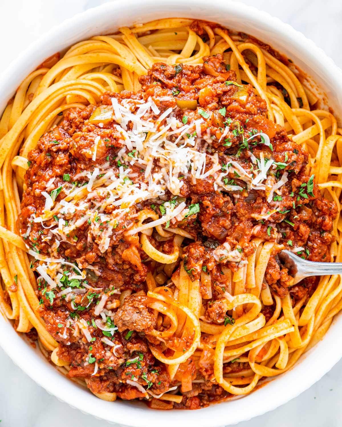 overhead shot of meat sauce over a bed of linguini with a fork twisting some pasta garnished with parmesan cheese and parsley
