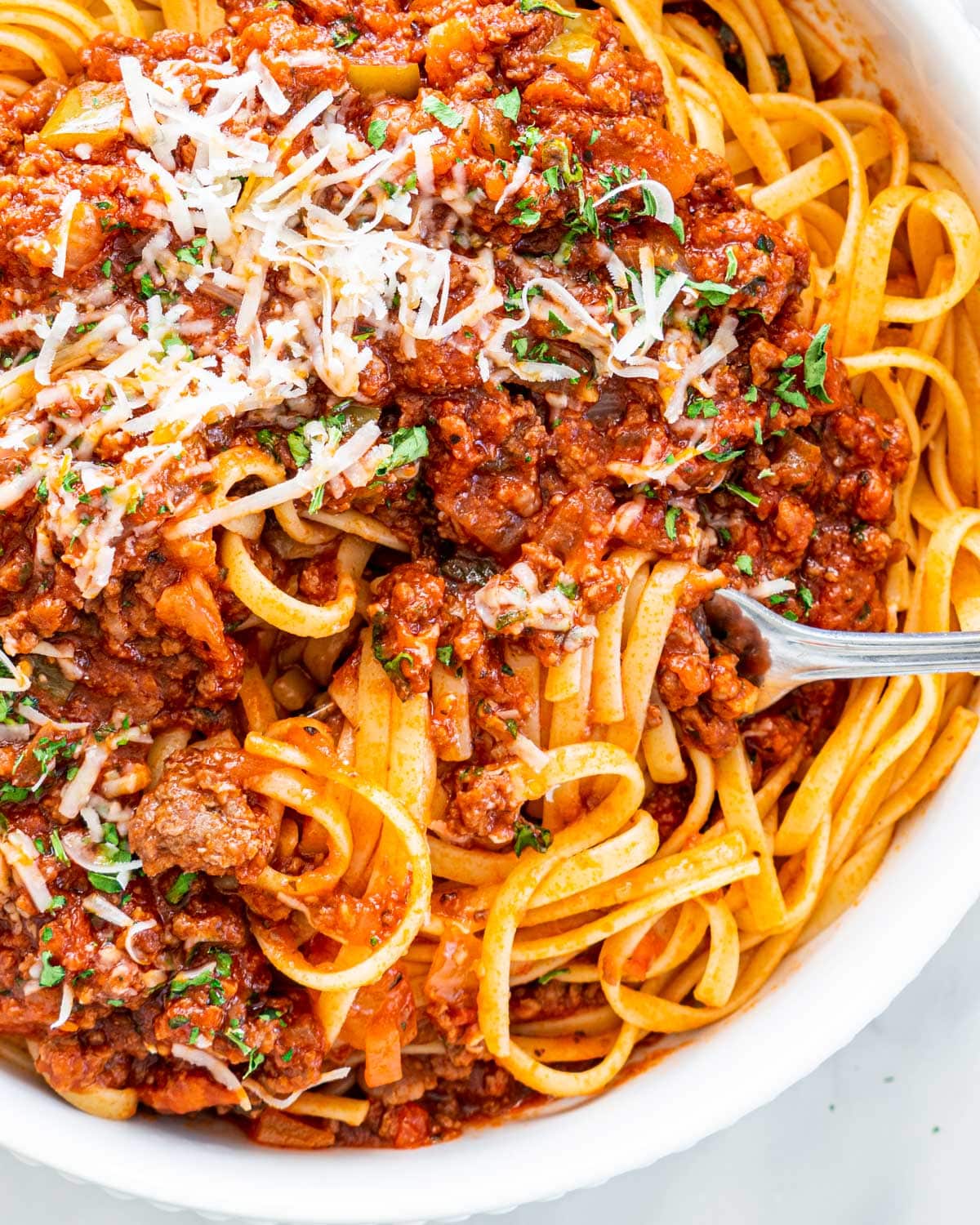 overhead closeup shot of meat sauce over a bed of linguini with a fork twisting some pasta garnished with parmesan cheese and parsley