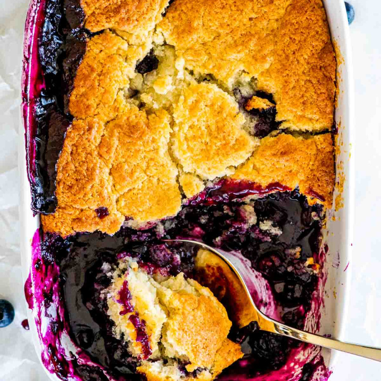 overhead shot of a blueberry cobbler in a baking dish with a a serving spoon taking a scoop out
