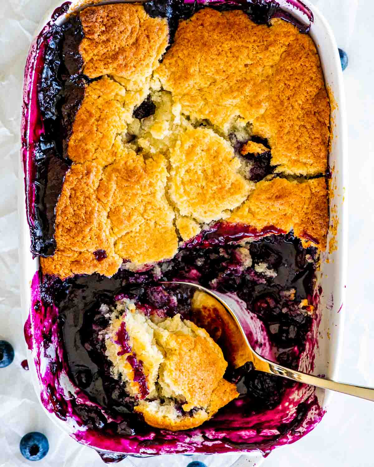 overhead shot of a blueberry cobbler in a baking dish with a a serving spoon taking a scoop out