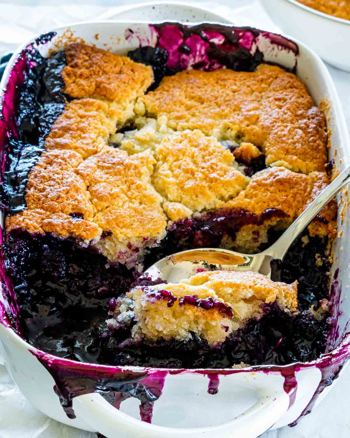 sideview shot of a blueberry cobbler in a baking dish with a a serving spoon taking a scoop out