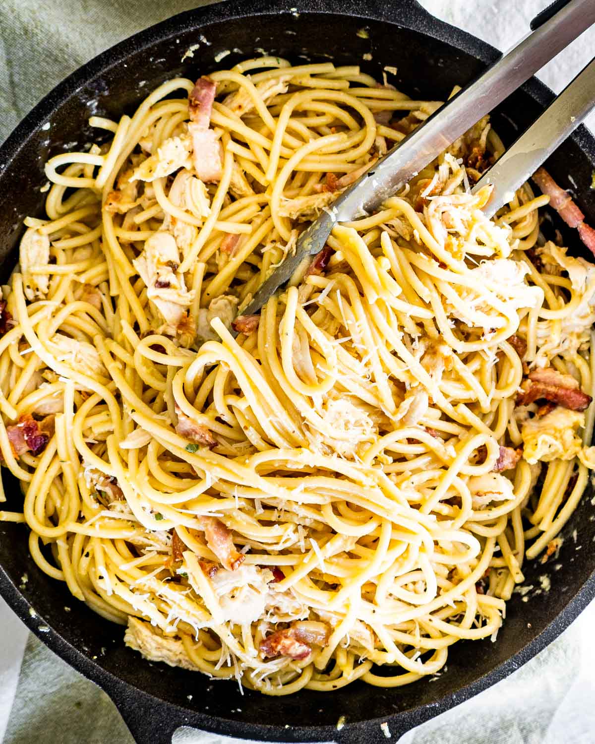 overhead shot of freshly made chicken carbonara in a black skillet with a pair of tongs in it