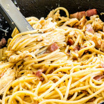 sideview shot of carbonara with chicken and bacon in a black skillet with a pair of tons in it