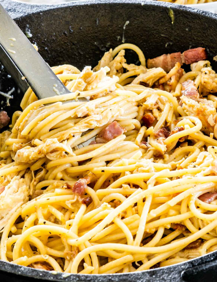 sideview shot of carbonara with chicken and bacon in a black skillet with a pair of tons in it